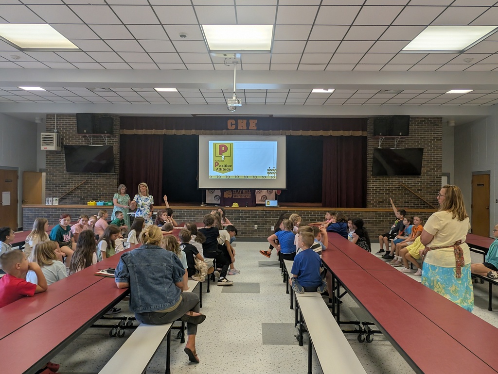 A large group of children seated at tables, watching a presentation on a screen at the front.