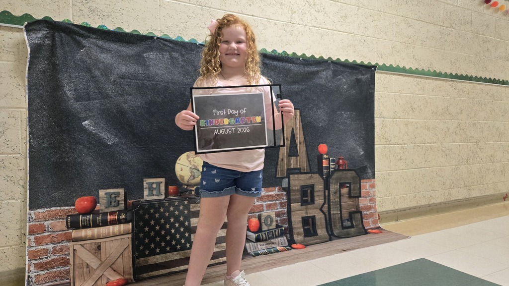 A young girl stands in a room with a black backdrop, holding an award. The backdrop has letters and a clock.