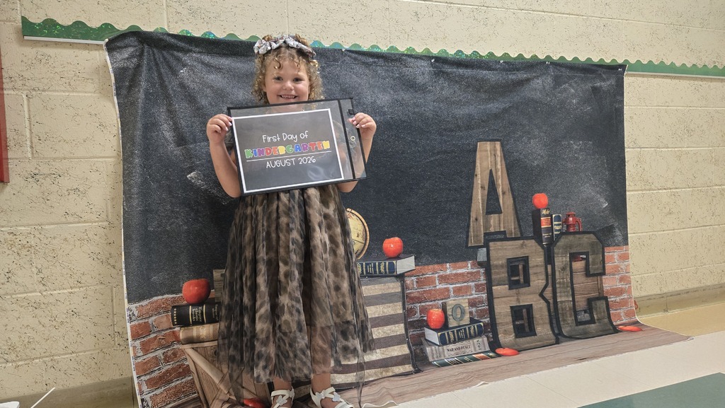 A young girl stands on a stage holding a framed certificate. Behind her is a brick wall with alphabet letters.