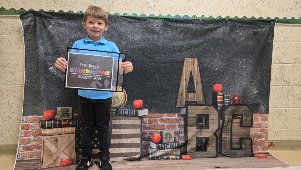 A child holds a certificate in front of a backdrop with a large "ABC" and a brick wall.