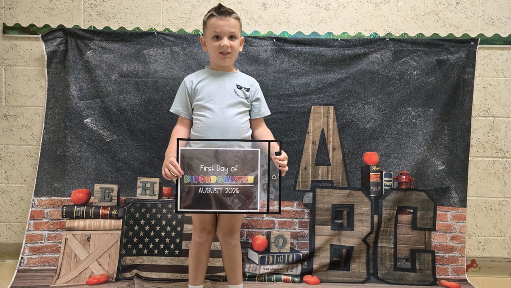 A young boy stands in front of a backdrop with letters and holds an award plaque.