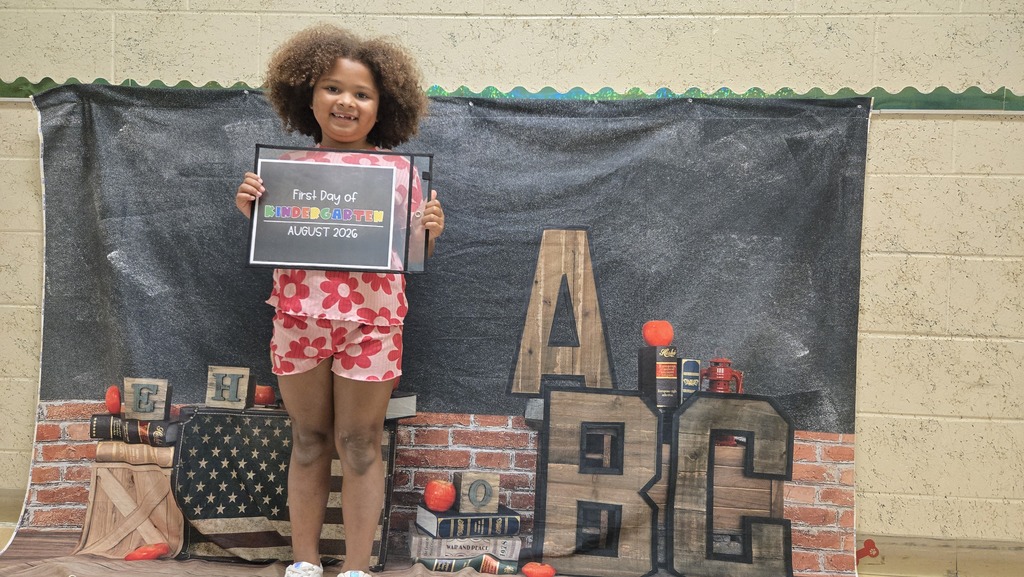 A girl in a floral dress and shorts holds a framed certificate in front of a chalkboard backdrop.