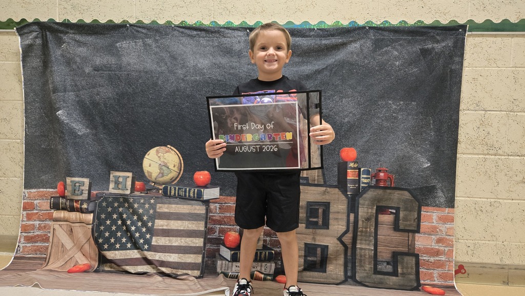 A child in black shorts and a shirt holds up a framed certificate in front of a decorated background.