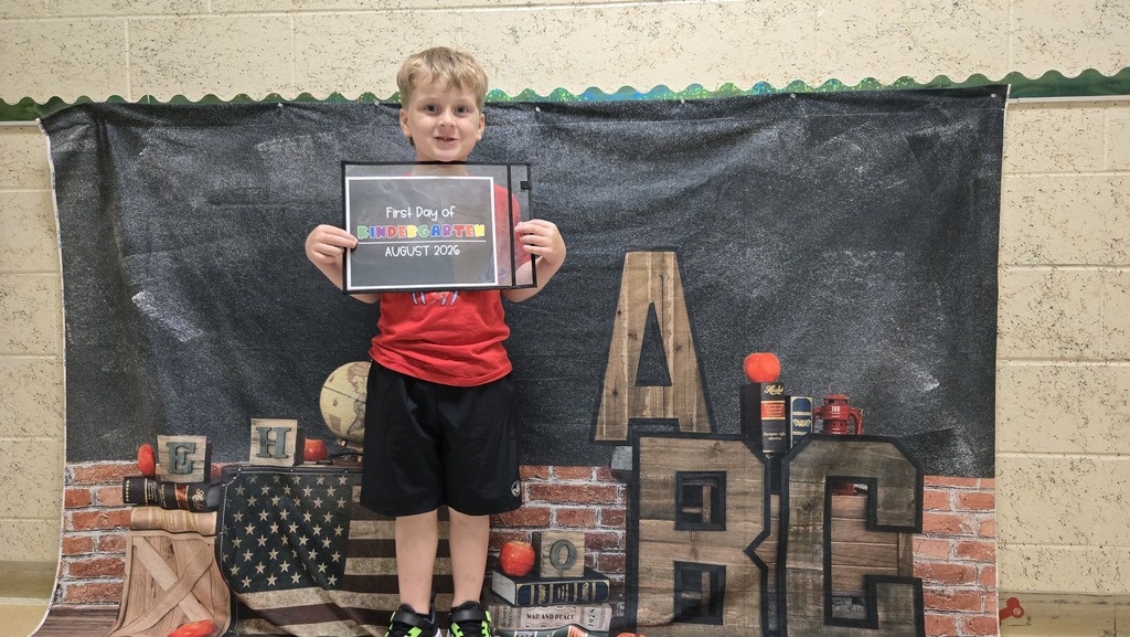 A young child stands, holding a framed award, before a banner featuring text and images.