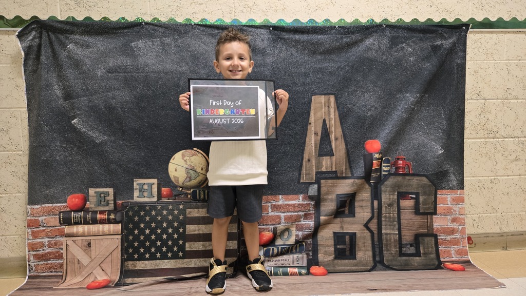 A child in front of a classroom backdrop holding a certificate. Behind the child is a wall with a brick design and educational props.