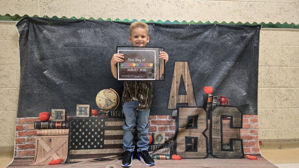 A child holds a framed certificate before a backdrop with bricks, a globe, and "ABC" letters.