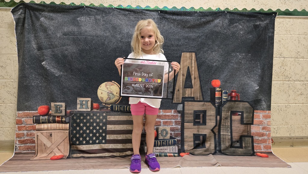 A young girl holding a framed certificate stands on a stage, with a backdrop of a blackboard and a brick wall.