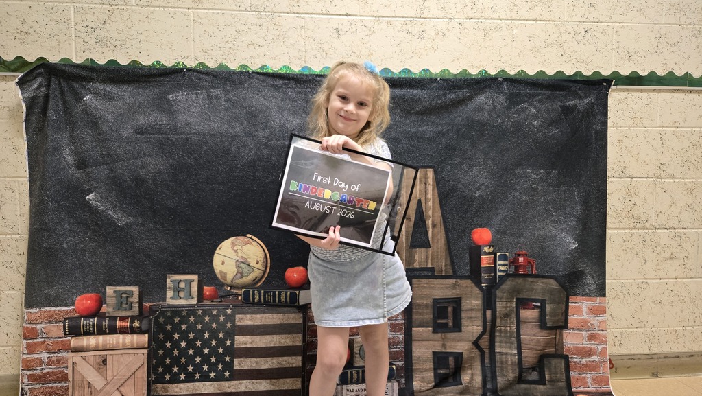 A young girl holds a framed certificate in front of a backdrop with a blackboard, globe, and American flag.