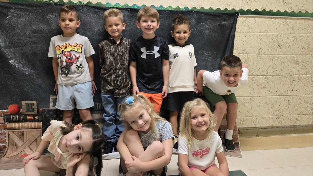 A group of children posing in a classroom, with one sitting on the floor, against a black backdrop.