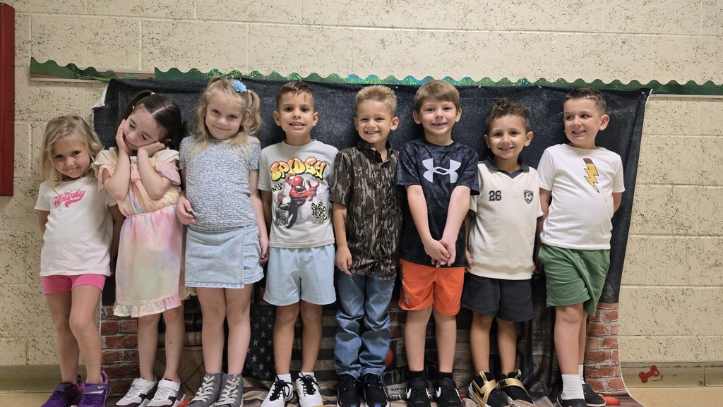 Nine young children in casual clothes standing in front of a brick wall with a black backdrop.