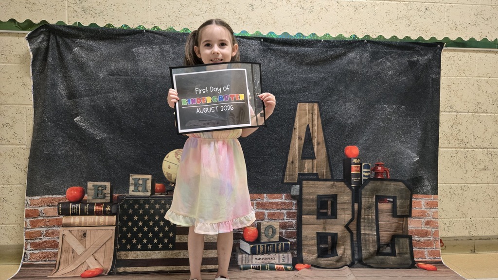 A young girl in a pink dress holds a certificate against a backdrop with letters ABC.