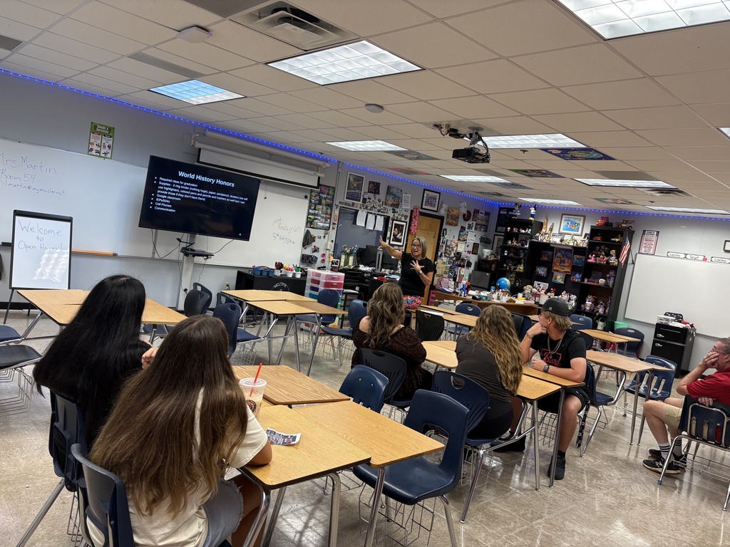 Classroom with students seated at desks; a teacher stands near the front, with a screen and whiteboard behind them.