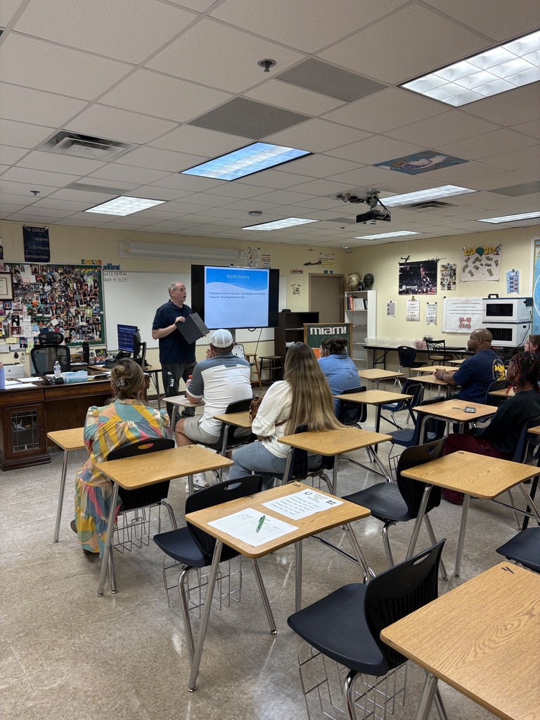 A classroom with white walls and a projector screen showing a presentation. A teacher stands at a podium. Students sit in rows of desks, listening.