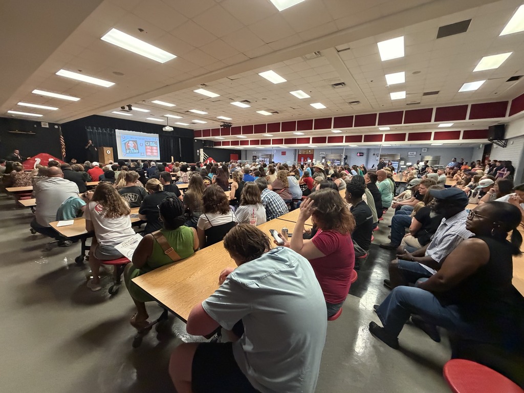 Many people sit at tables in a large room with a projector screen at the front.