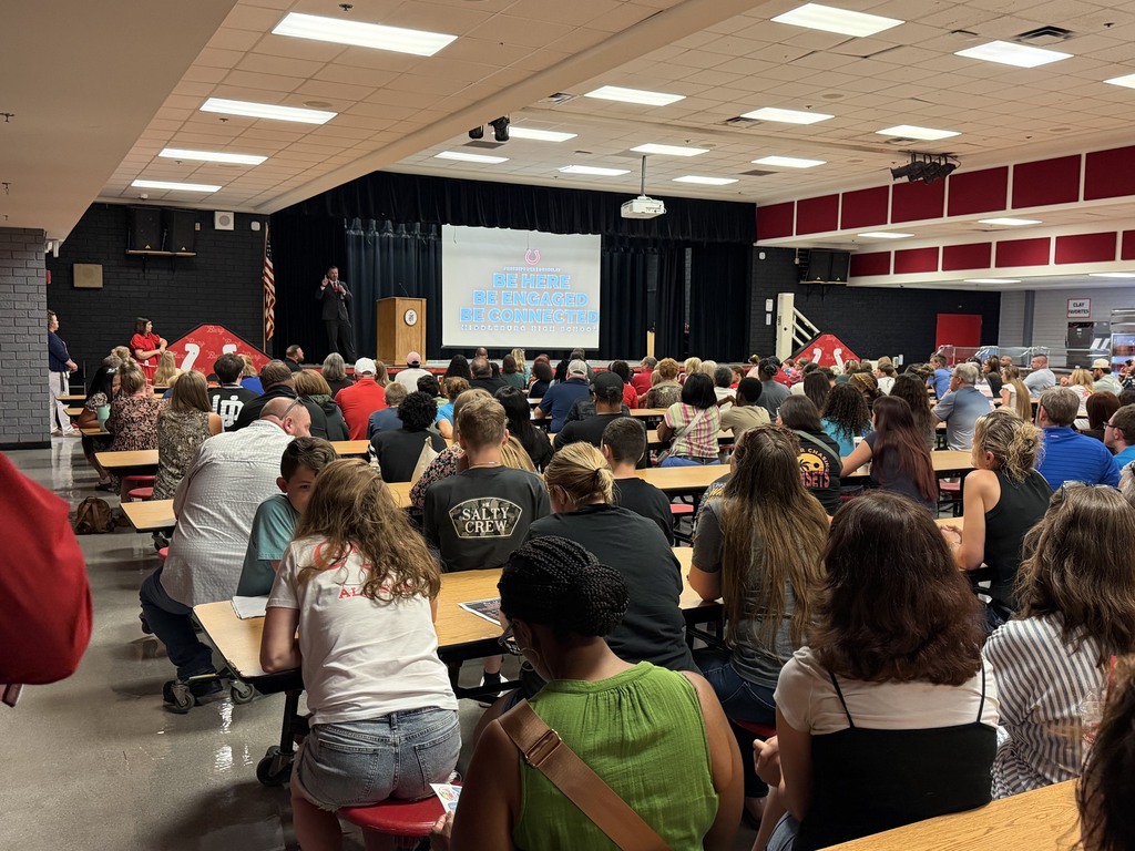 A large room with people seated at tables, listening to a presentation on a projector screen.