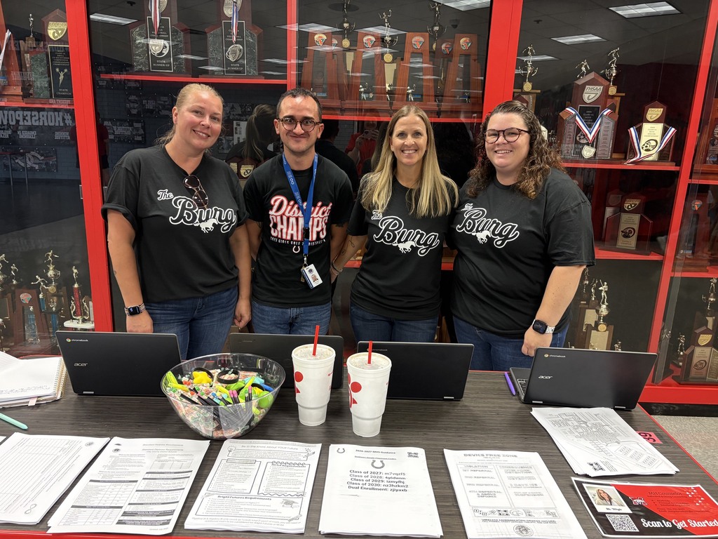 Four people in matching shirts stand behind a table with laptops and papers, with shelves of trophies in the background.