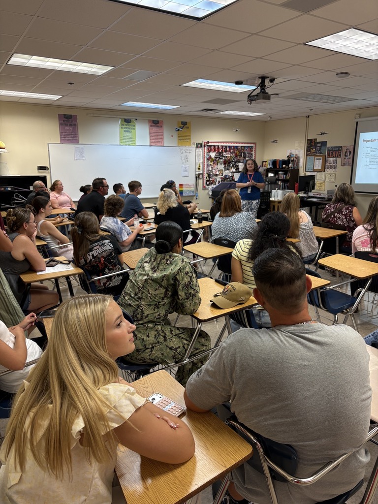 A classroom with desks and chairs. A teacher at the front is addressing a group of students.