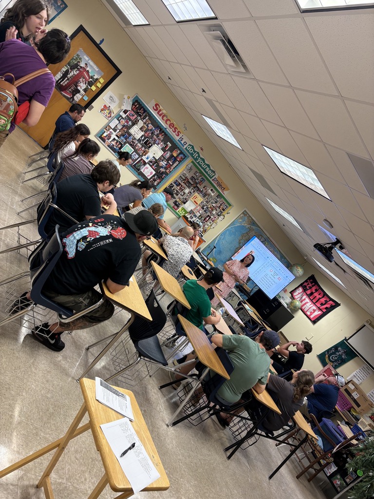 A classroom with students seated at desks, a teacher standing at the front, and a projector screen displaying information.