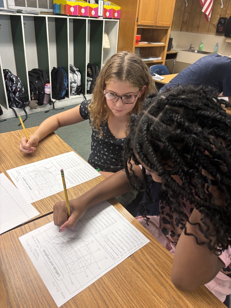 Two girls seated at a desk, each holding a pencil and writing on white papers. A classroom with shelves and a flag in the background.