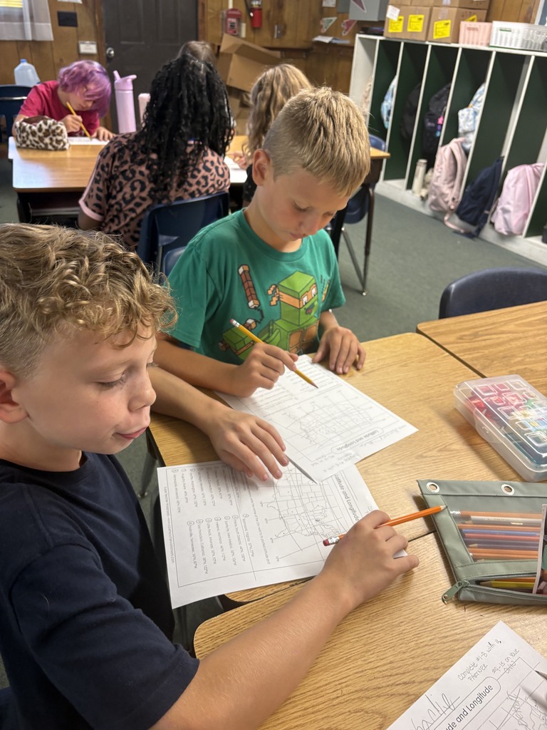 Kids seated at a desk in a classroom, one using a pencil to fill out a worksheet.