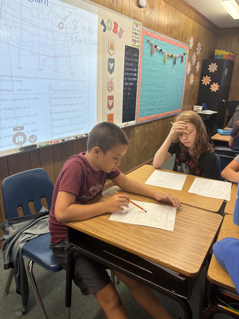 Two children seated at desks, engaged in writing. Behind them, a classroom with a whiteboard and decorative posters.