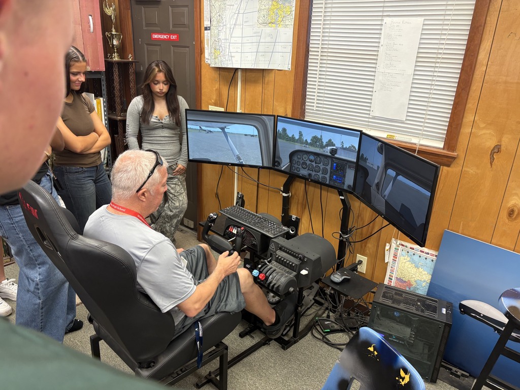 Four people are in a room with wood paneled walls. Three people are looking at a man using a flight simulator.