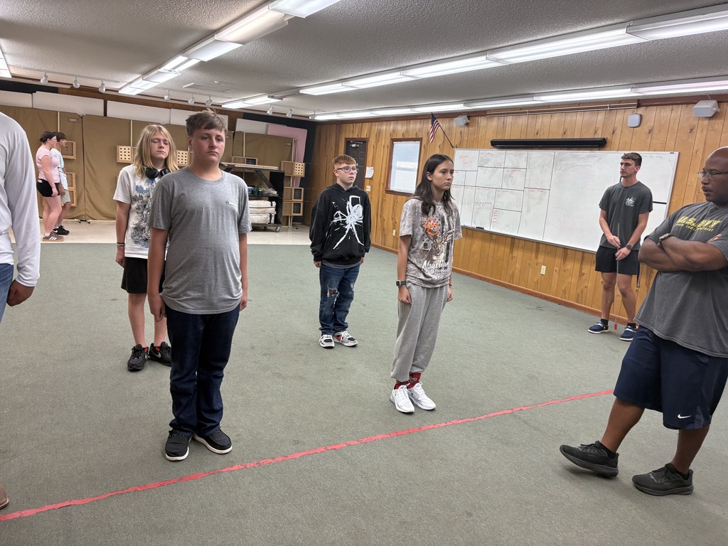 Six individuals, including young adults, stand in a room with white walls and wooden paneling. Two people cross their arms, while others appear to listen.