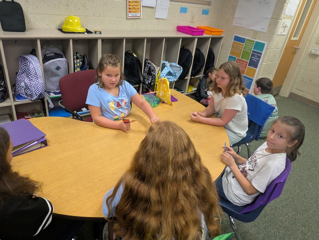 A group of children are sitting around a wooden table, facing each other and talking. Some wear backpacks.