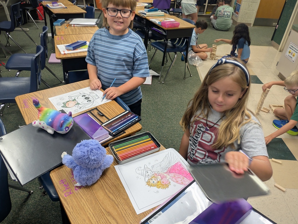 Children seated at desks in a classroom, one using a tablet, the other with coloring tools and toys.