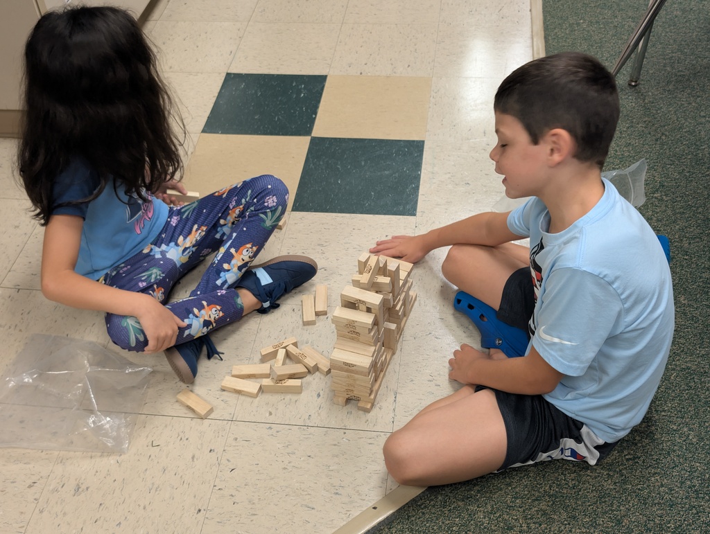 A child and a girl play with wooden blocks on a floor with a checkered pattern.