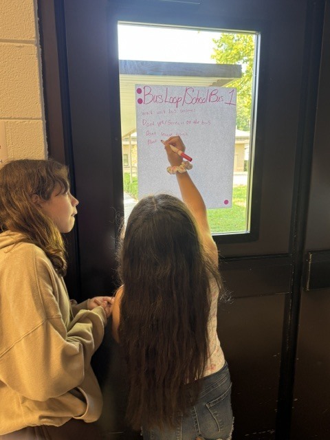 Two young people write on a sign next to a window, with one holding a pen and the other looking on.