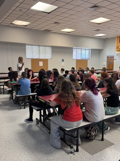 Room with tables and benches; a woman stands and addresses seated students in a cafeteria setting.