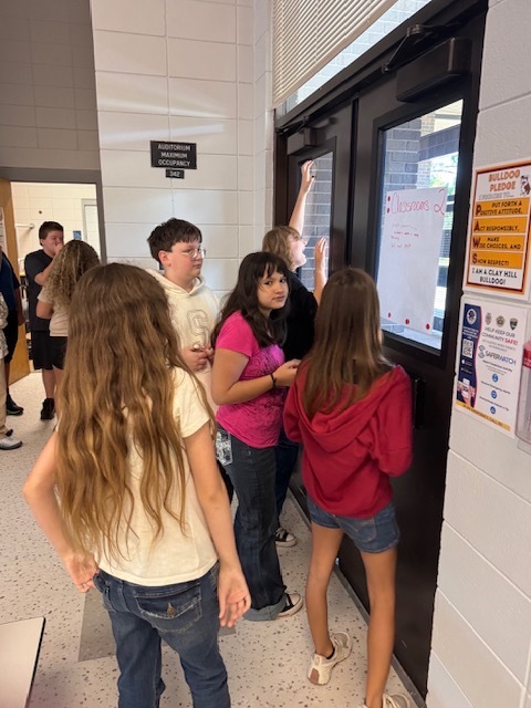 A group of students stands in front of a door with a screen and writing, discussing.