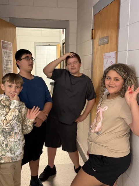 Four children in a hallway, two boys and two girls, standing together and smiling, one girl leaning against a wall.