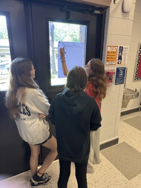 Three students stand by a door, one writing on a board, another looking out the window.
