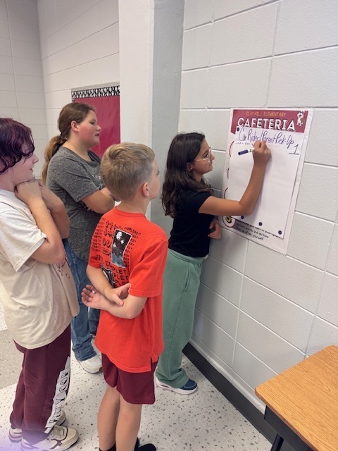 A group of four young children in a room. They are looking at and writing on a large white board.