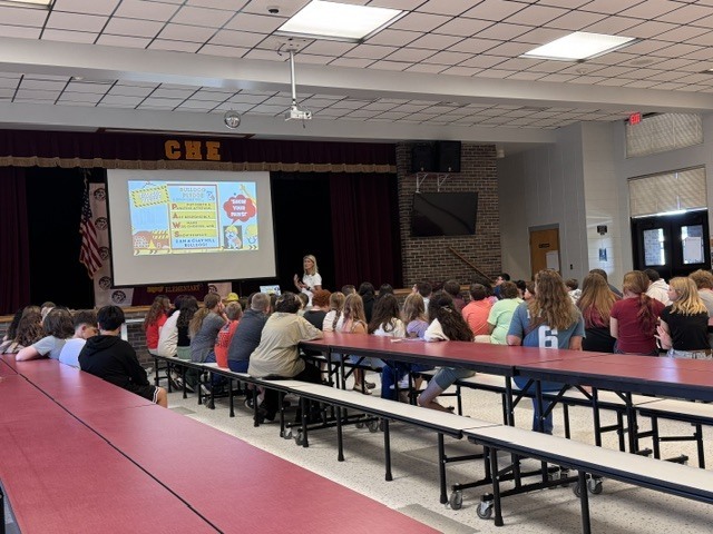 A classroom setting with a woman at a podium, screen, and students seated at tables.