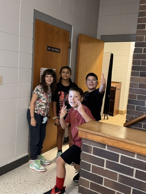 Four children stand in a hallway with two open doors. One boy smiles. A brick wall is on the right.