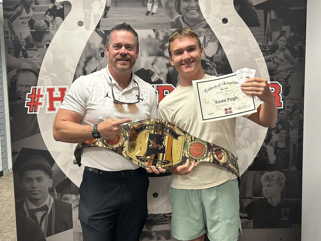 Two men stand next to each other in front of a decorative wall. One holds a championship belt, and the other holds a certificate.
