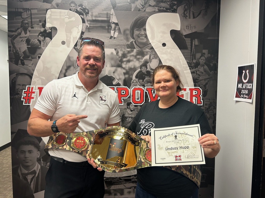 A man and a woman stand in front of a large poster, holding a championship belt and certificate.