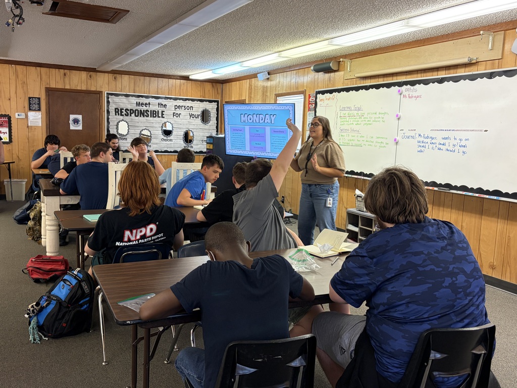 A classroom with wooden walls; students seated in front of desks and a teacher standing before a screen.