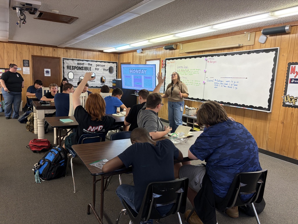 People seated in chairs around tables in a classroom. A woman stands near a whiteboard and presents to a group.