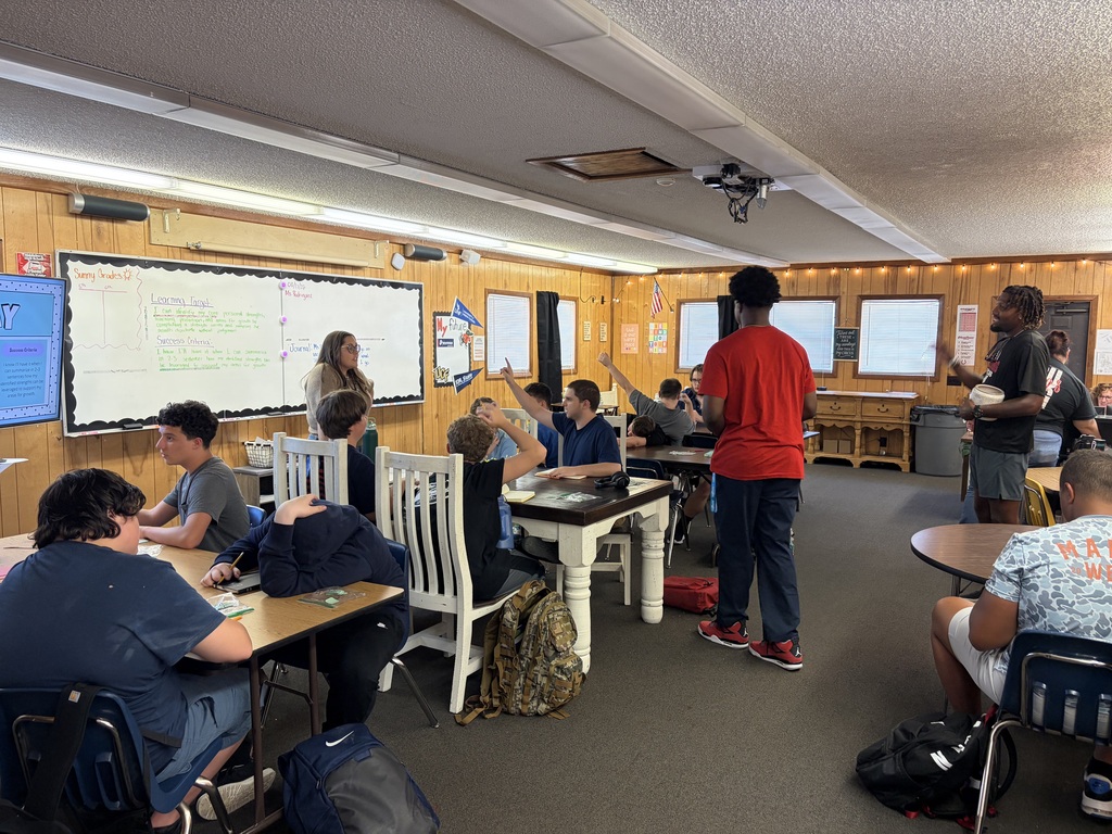 A classroom with wooden walls. Students sit at desks, one standing at a whiteboard. Bags and backpacks are on the floor.