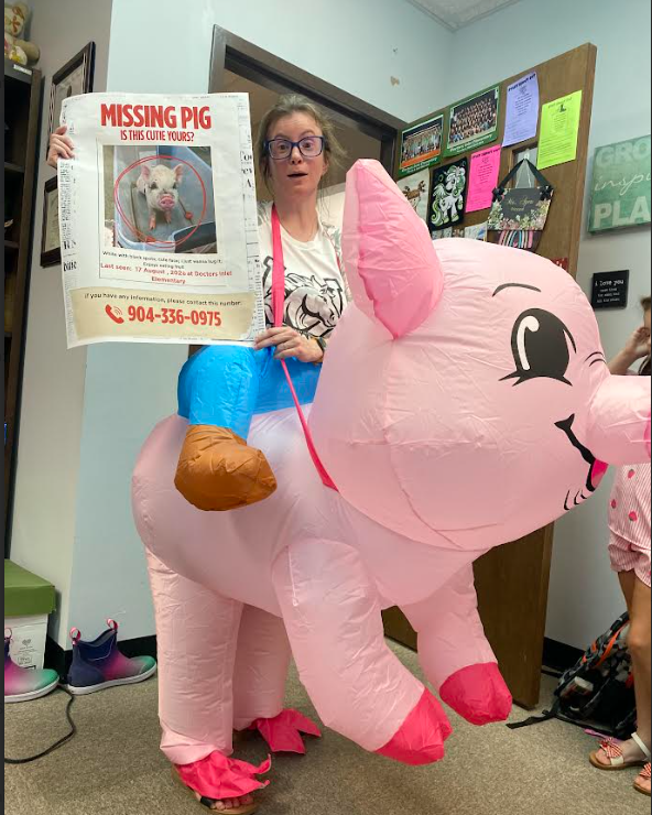 A person in a pink inflatable pig costume holds a missing pig poster in a room.