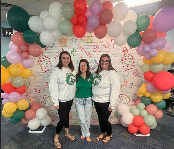 Three people stand before a colorful balloon arch against a white wall with drawings.