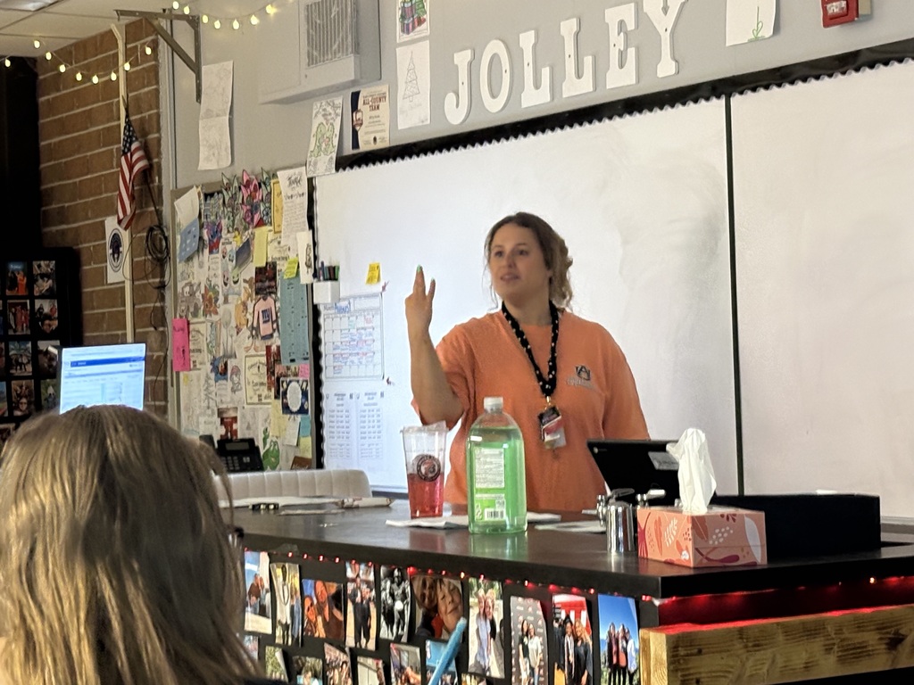 A teacher in an orange shirt is speaking in front of a whiteboard. A student sits before the teacher.