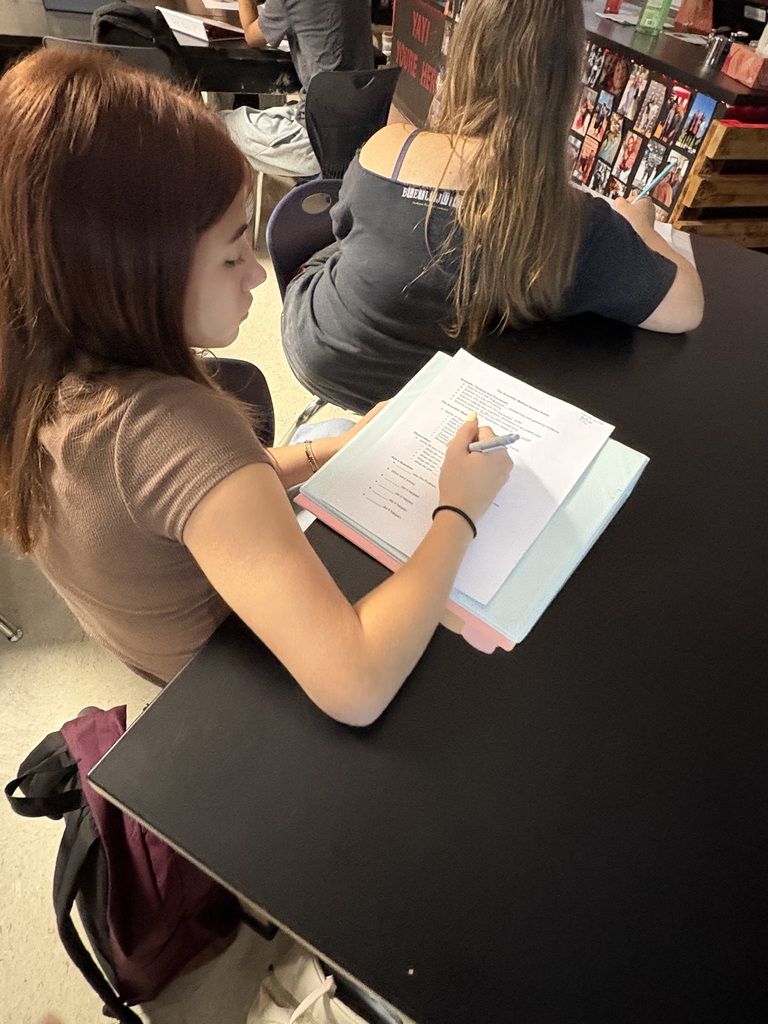 Two students work at a desk, one writing with a pen on paper, the other seated nearby.