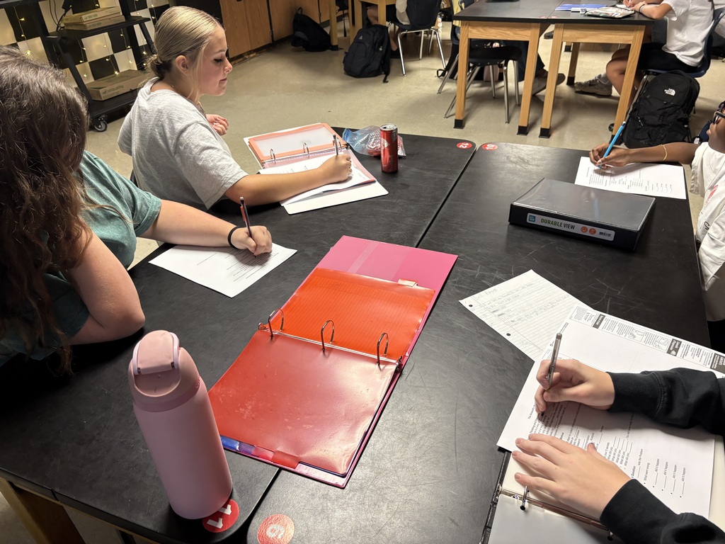 Four people at desks in a classroom. Papers, binders, and pens are on the desks.