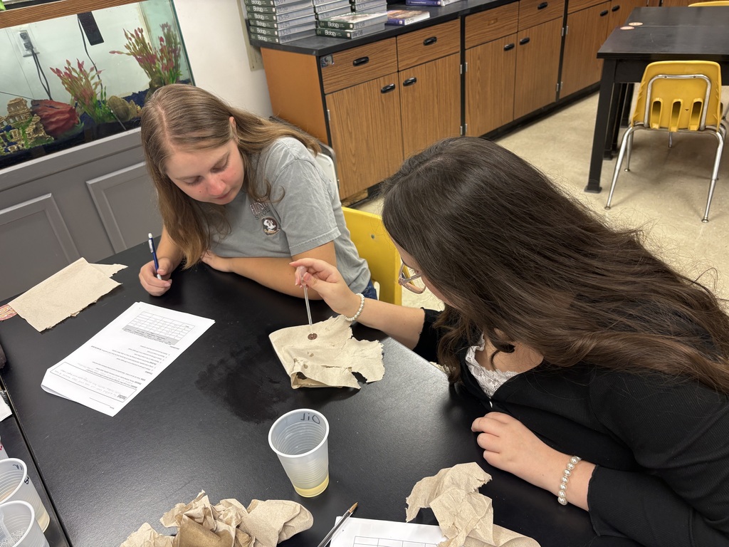 Two women are working at a table. One holds a pen, and the other holds a cloth.