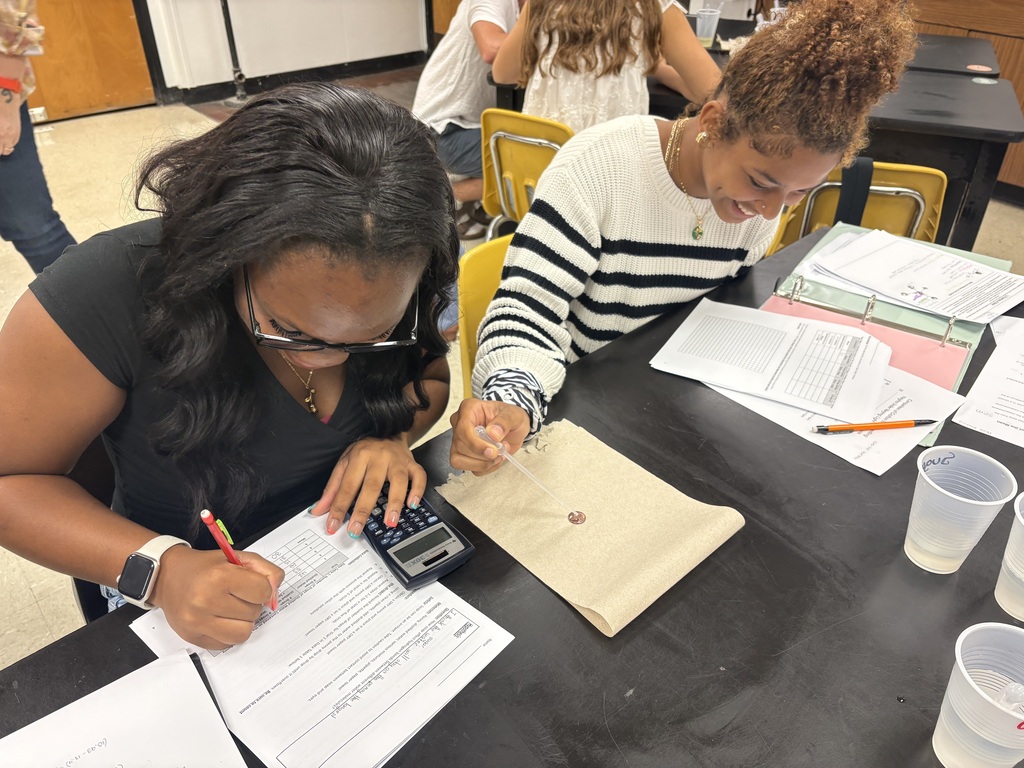 Two individuals at a table, one using a calculator, the other writing on paper. Papers, pens, and a calculator are on the table.